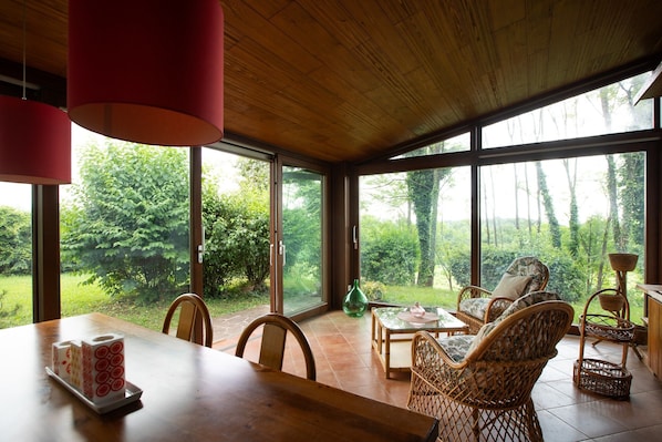 A dining area with a wooden table, wicker chairs, and a view of greenery through large windows.