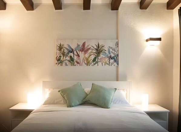 Un lit avec deux oreillers, une table de chevet, une lampe fixée au mur et une œuvre d’art florale au-dessus du lit.