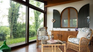 A room with wicker furniture, a glass-top table, and a view of greenery through large windows.