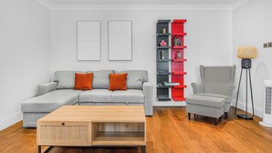 A modern living room with a grey sectional sofa, a wooden coffee table, a patterned armchair, a red and black shelving unit, and a floor lamp. - A Scholar s Gallery - Bloomsbury - by Frankie Says (London)