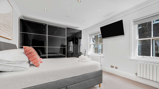 A modern bedroom with a large bed, a walk-in closet, a flat-screen TV, and a window with a view of a brick building. - A Scholar s Gallery - Bloomsbury - by Frankie Says (London)