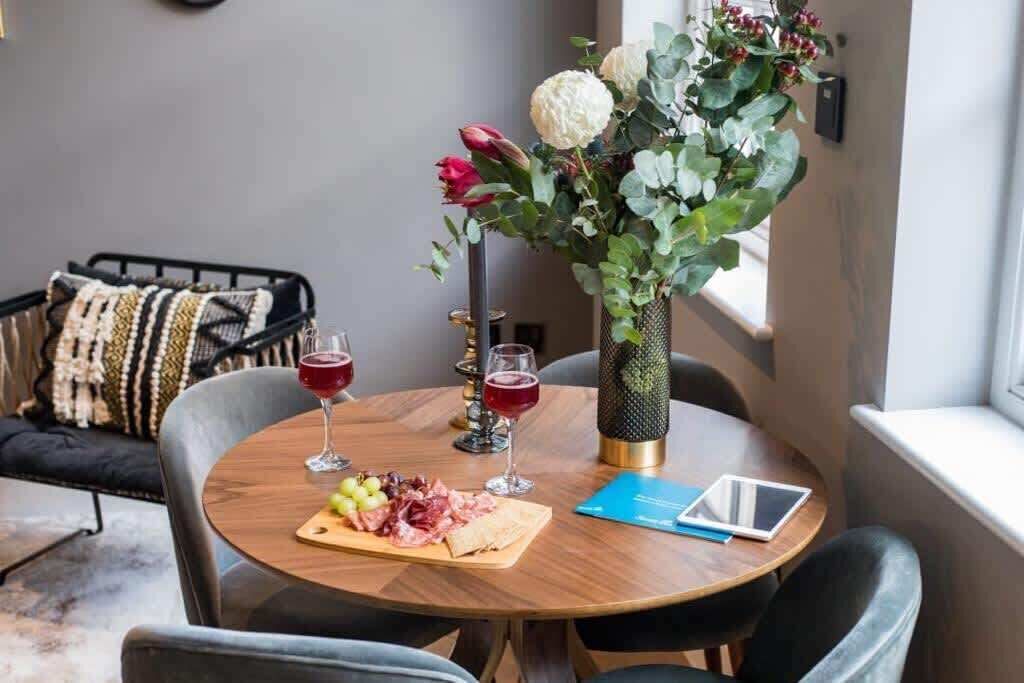 Taula de fusta rodona amb un got de vi negre, un plat de formatge i raïm, i una tauleta. Sobre la taula hi ha un ram de flors i vegetació.