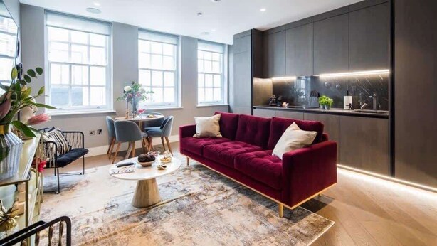 A modern living room with a red sofa, a coffee table, and a dining area with chairs. There are large windows providing natural light and a kitchen area with dark cabinetry and a countertop. - The Avicii Signature - Soho - by Frankie Says (London)