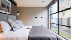 A modern hotel room with a large bed, a view of the city through large windows, and a painting on the wall. - The Avicii Signature - Soho - by Frankie Says (London)