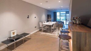 A modern dining area with a long table and bar stools, a bench, and a view of the outdoors through large windows. - Monochrome Majesty - Soho - by Frankie Says (London)
