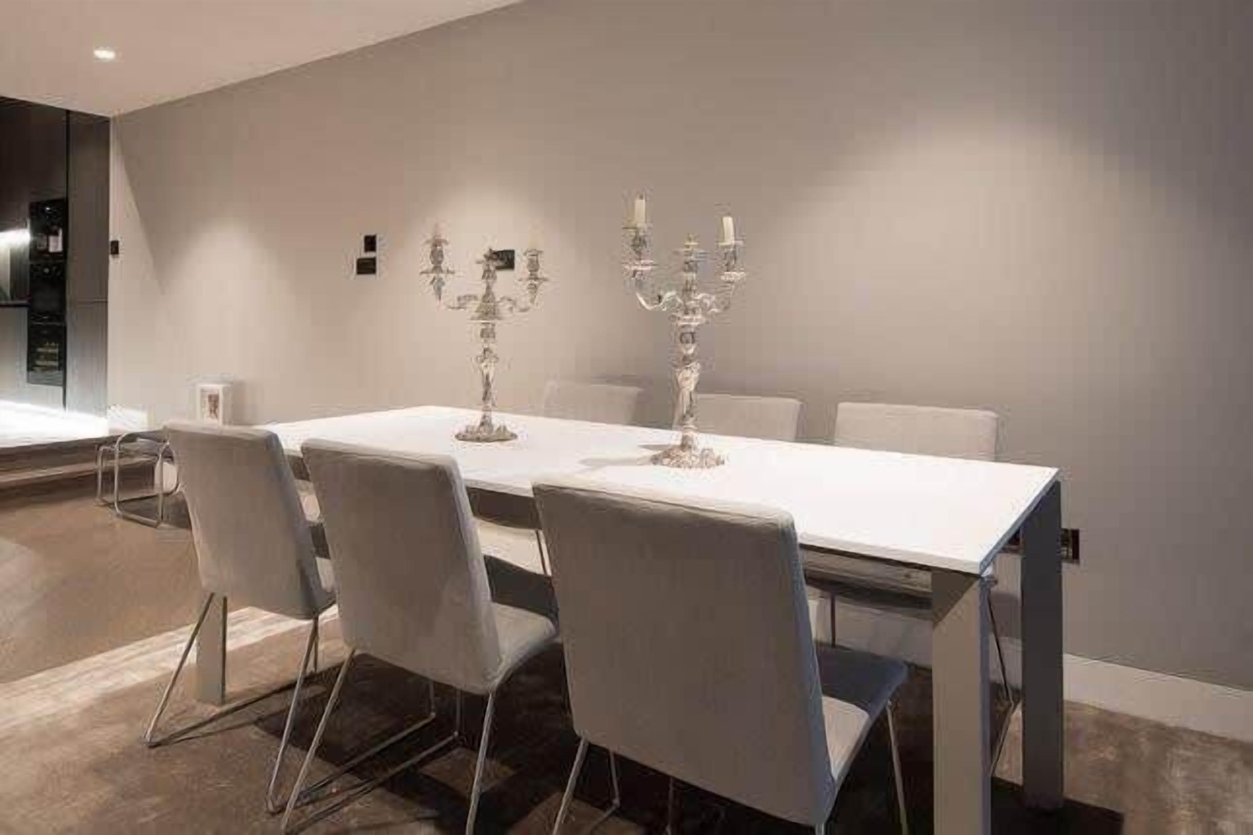 A modern dining area with a white table and grey chairs, two candelabras on the wall, and a visible kitchen area to the left.