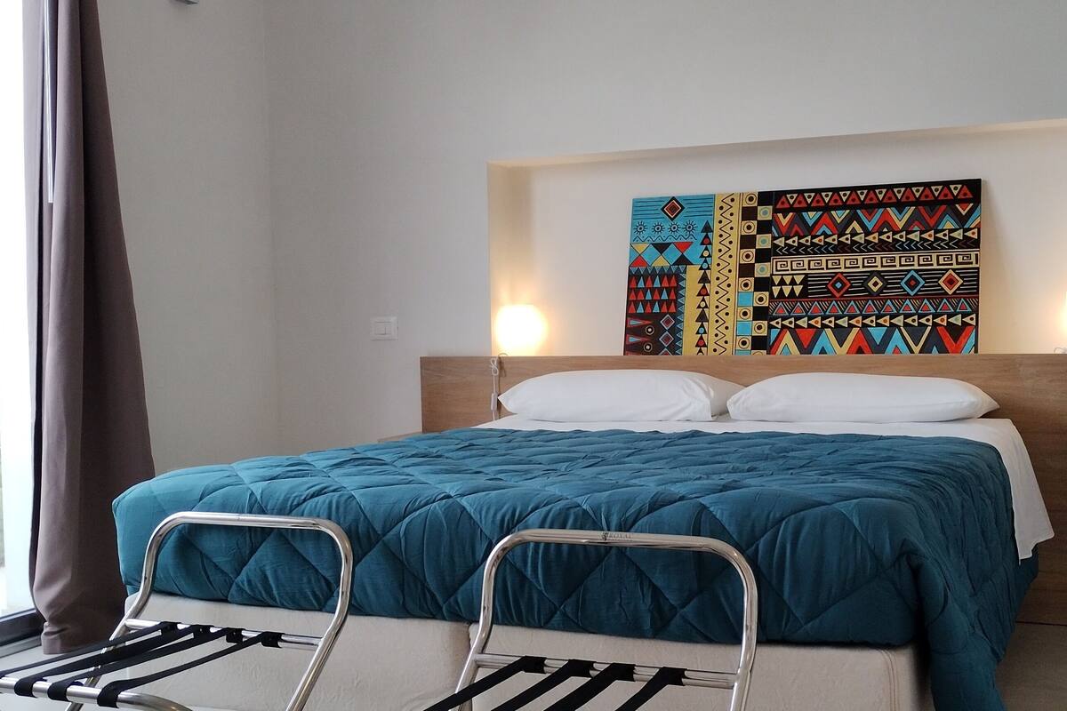 A bedroom with a bed, a headboard, a wall-mounted artwork, and a window with curtains.