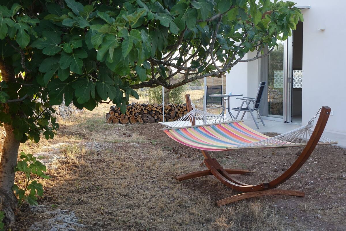A hammock under a tree, a pile of firewood, and a patio area with a table and chairs.