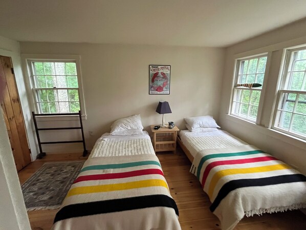 Una habitación con dos camas, una mesita de noche con lámpara, un cartel en la pared y una ventana con vistas a árboles.