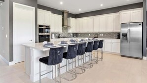 A modern kitchen with white cabinets, a stainless steel refrigerator, and a bar area with four stools.