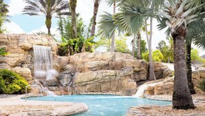 A pool area with a waterfall, palm trees, and a rocky backdrop.