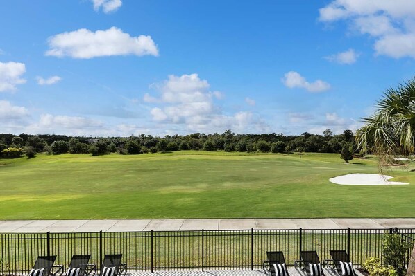 A golf course with a clear sky, a well-maintained green, and a fence with chairs in the foreground.