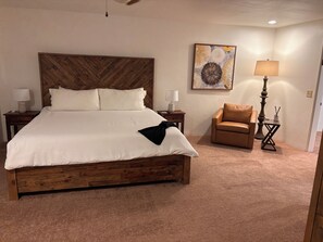 A hotel room with a large bed, two bedside tables with lamps, a brown leather armchair, a small side table with a lamp, and a wall-mounted artwork.
