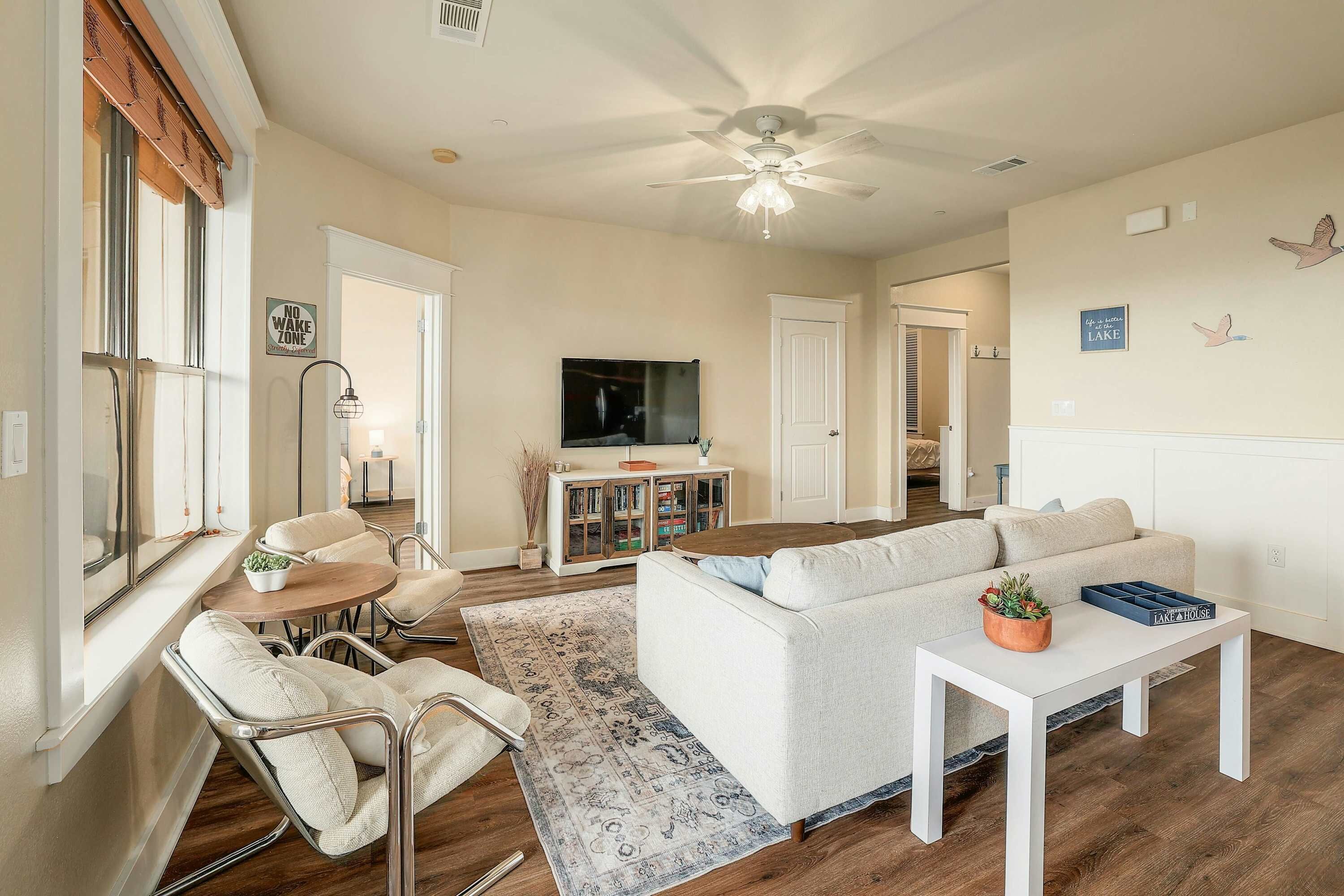 A living room with a white sofa, a coffee table, and a TV. There is a ceiling fan, a rug, and a window with blinds.