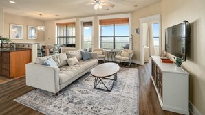 Un espacio habitable amplio con un sofá, una mesa de centro y un televisor. Se aprecia un balcón con vistas, y una cocina al fondo.