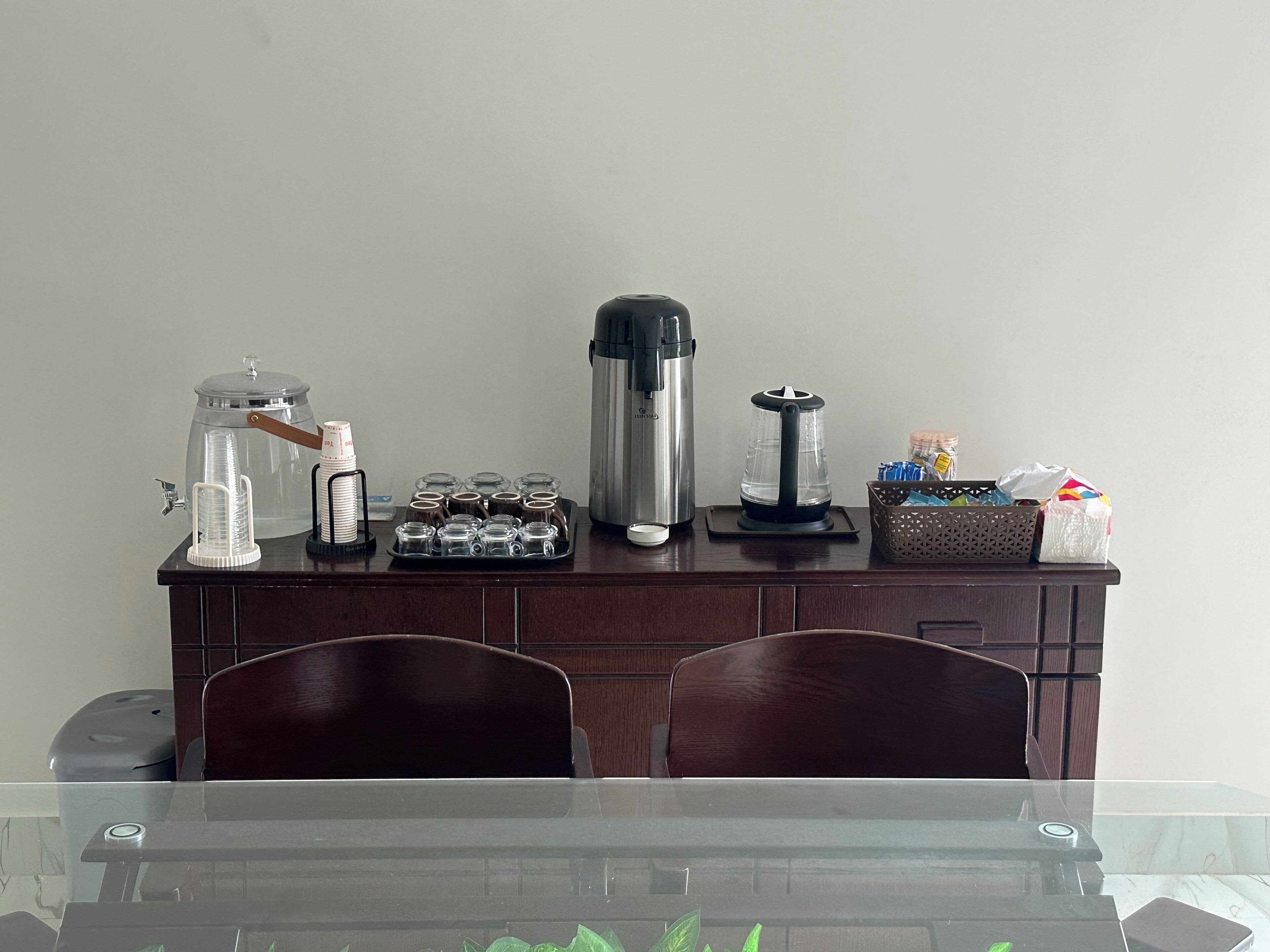 A dining area with a glass table, wooden chairs, a coffee station with a coffee maker, a water dispenser, and a basket of snacks.
