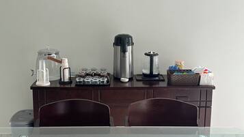 A dining area with a glass table, wooden chairs, a coffee station with a coffee maker, a water dispenser, and a basket of snacks.