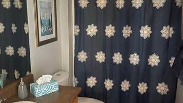 Uma casa de banho com sanita branca, toucador de madeira com lavatório, quadro emoldurado na parede e cortina de chuveiro com padrão floral.