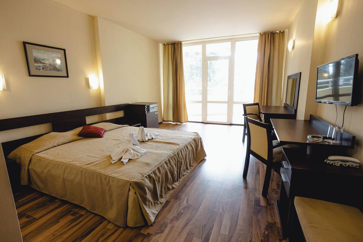 A hotel room with a large bed, a desk with a chair, a television, and a window with curtains.