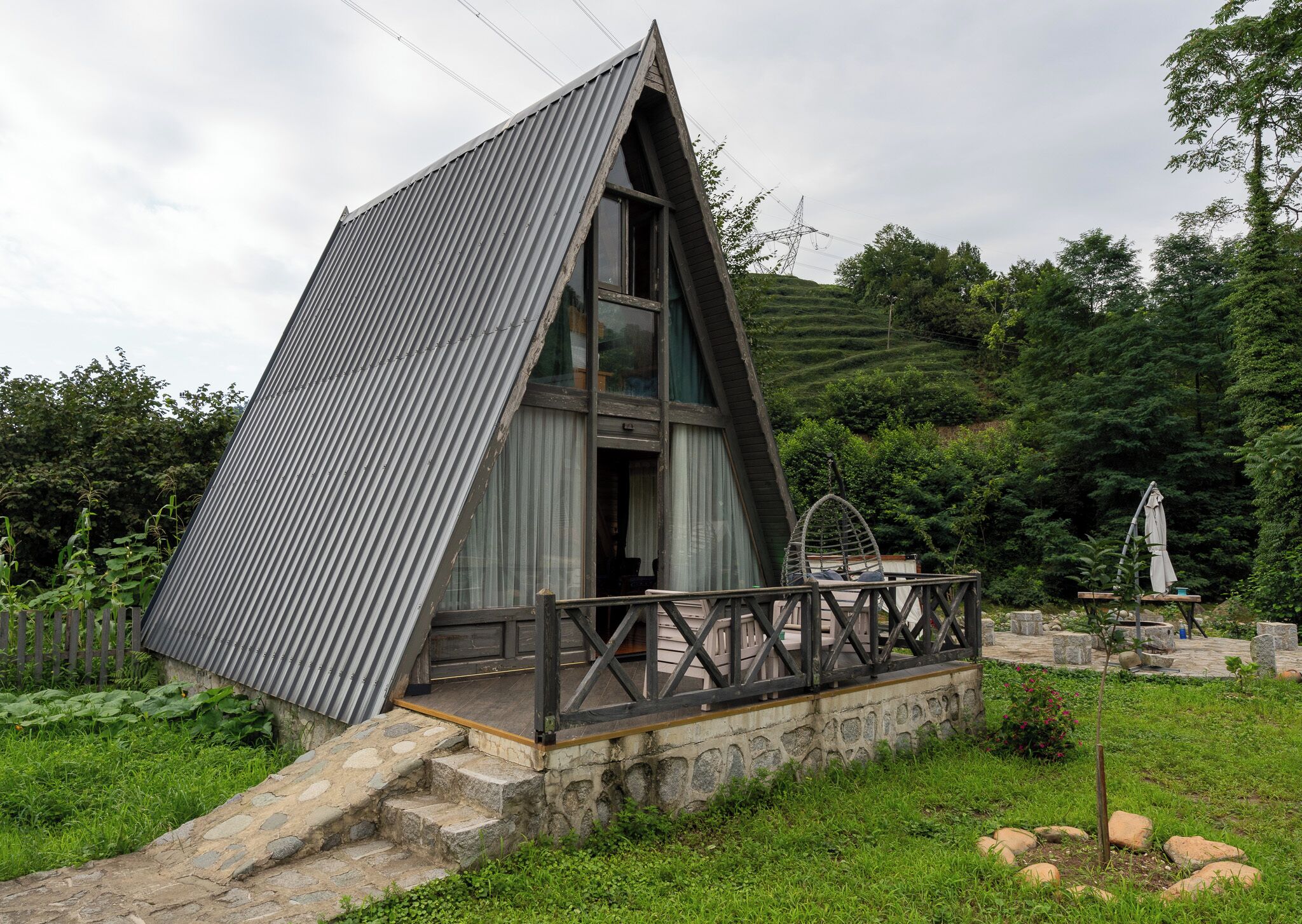 Une cabane triangulaire en bois avec une porte en verre, entourée d’un chemin de pierre et d’une rambarde en métal.
