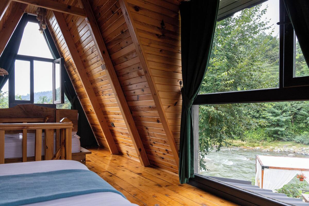 A room with wooden sloped ceilings, a bed, a bench, and a large window overlooking a river.