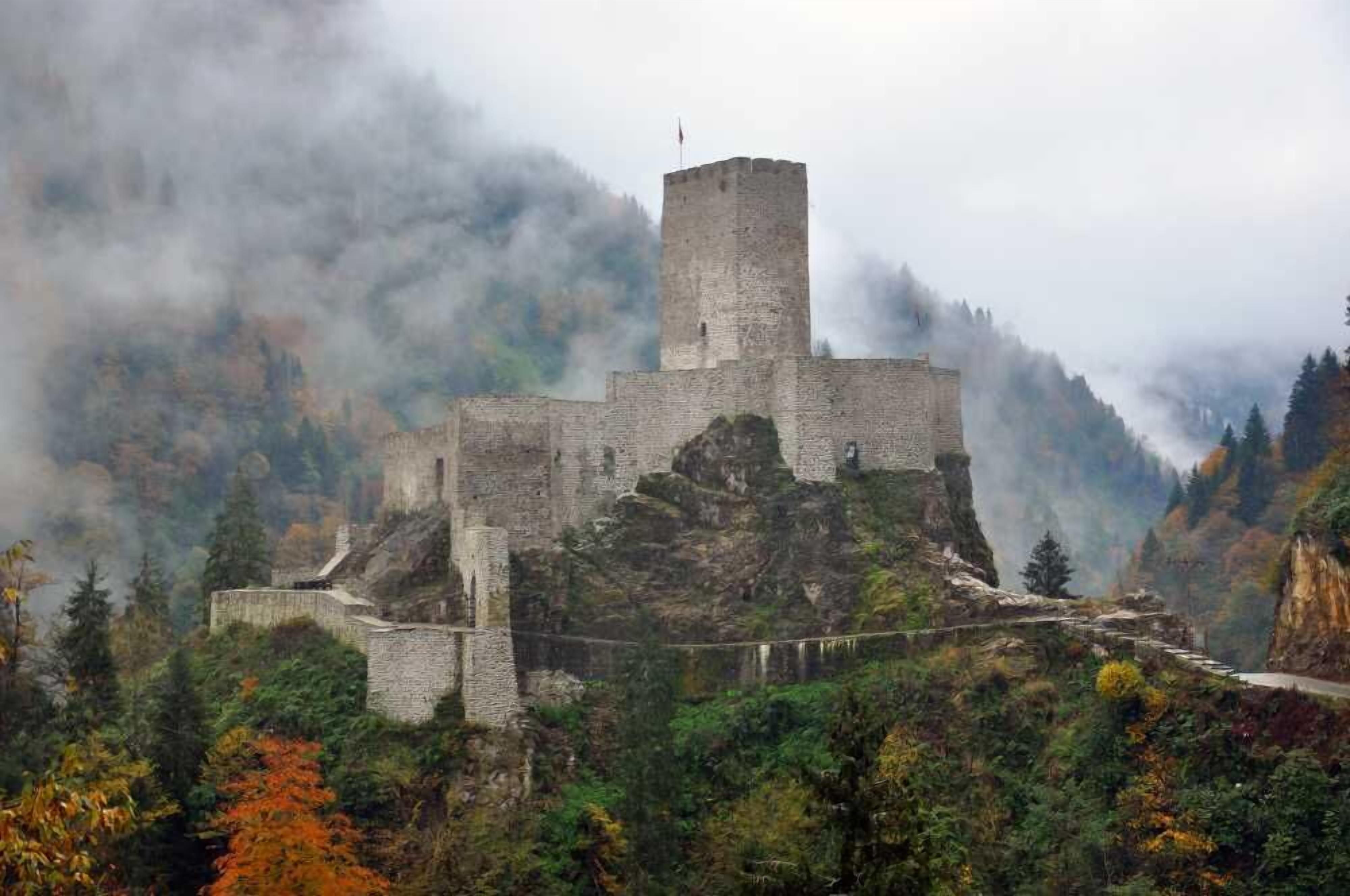 Een historisch stenen kasteel hooggelegen op een berg, omringd door herfstkleuren en nevel.
