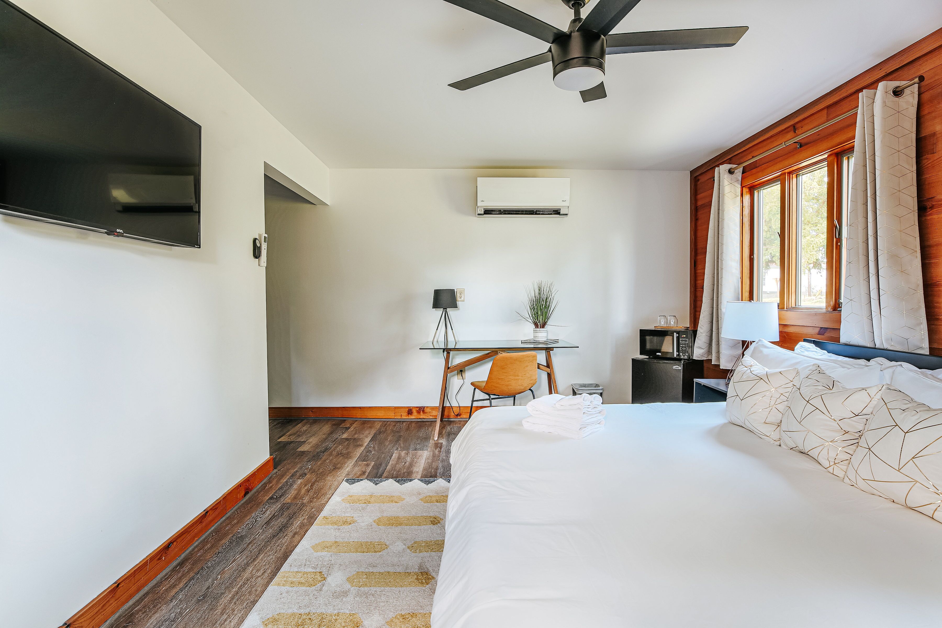 A hotel room with a bed, a desk with a chair, a ceiling fan, an air conditioning unit, and a window with curtains.