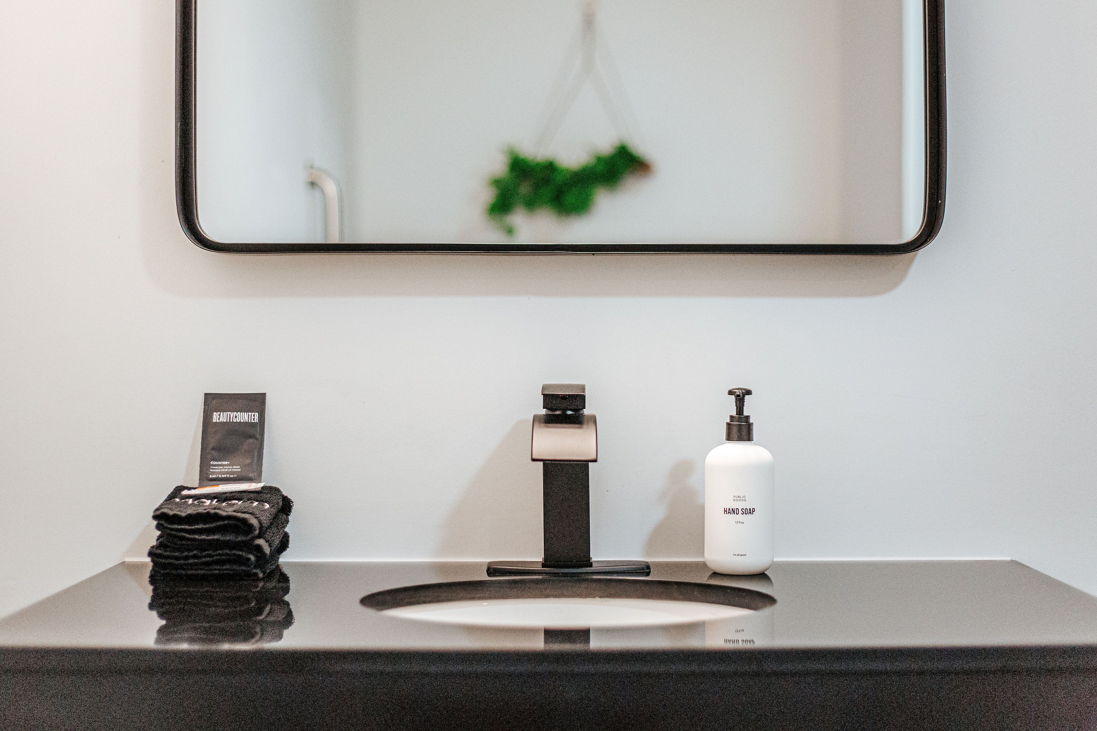 A bathroom with a black countertop, a sink with a modern faucet, a soap dispenser, and a mirror with a green plant reflection.