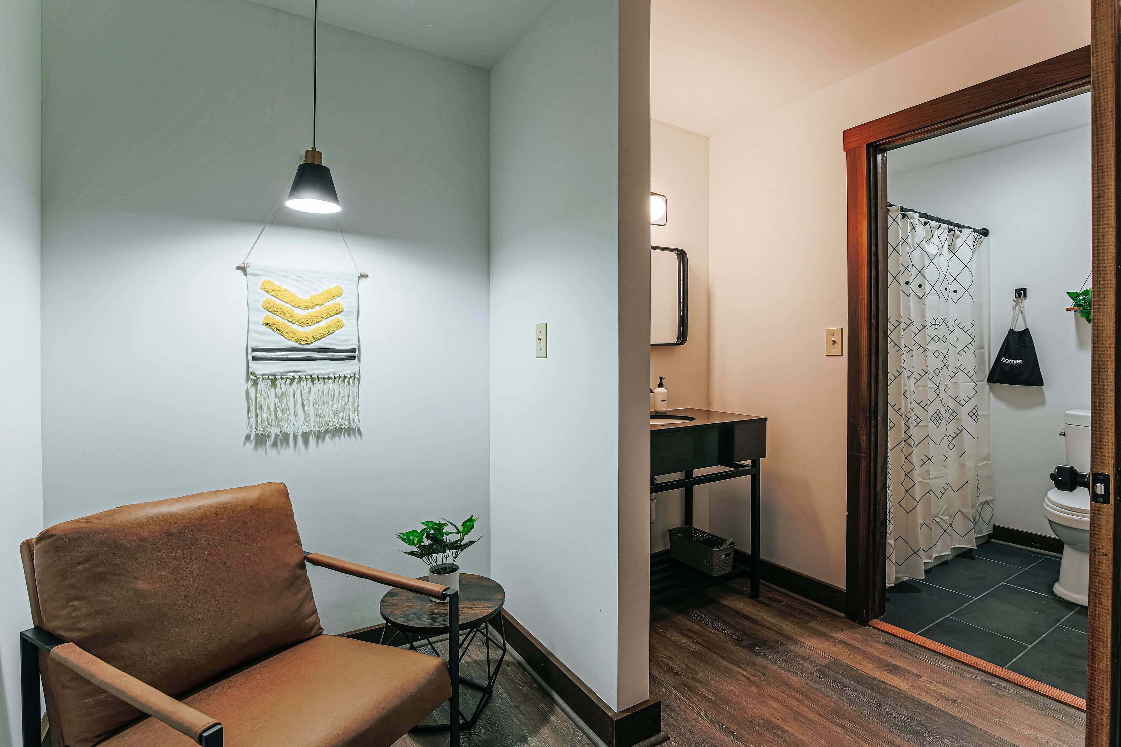 Una habitación moderna con un sillón de cuero marrón, una pequeña mesa redonda con una planta, una lámpara de pared y un espejo que refleja un baño con cortina de ducha estampada.