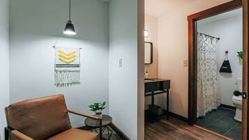 Una habitación moderna con un sillón de cuero marrón, una pequeña mesa redonda con una planta, una lámpara de pared y un espejo que refleja un baño con cortina de ducha estampada.