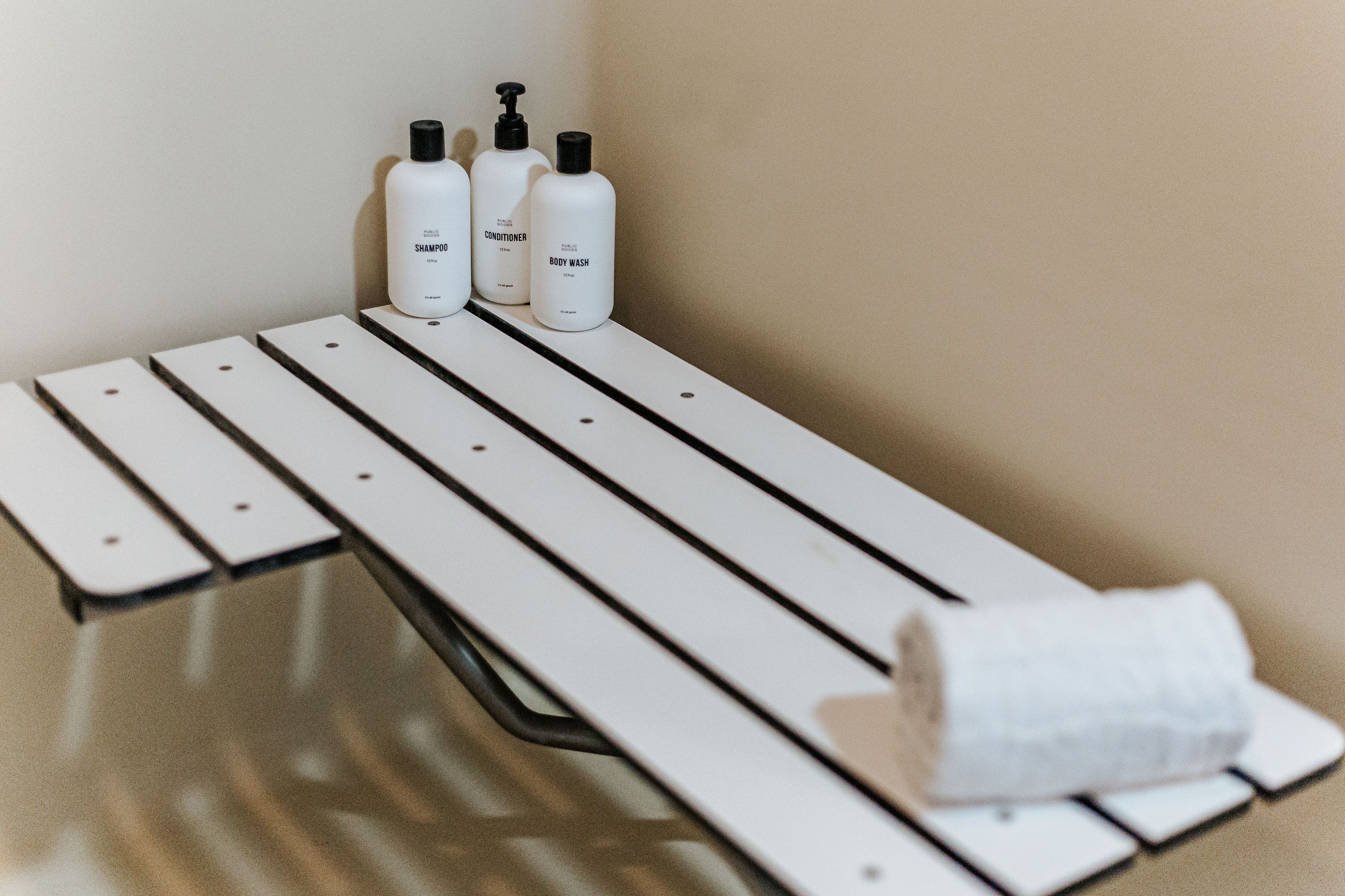 A bathroom with a white bench, three bottles of toiletries, and a rolled-up towel.