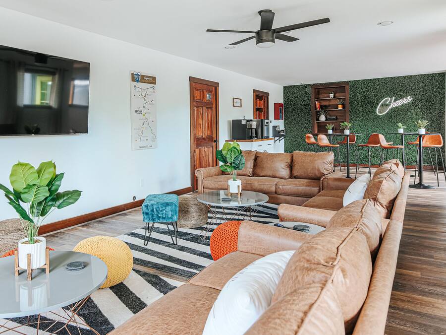 Uma sala de estar moderna com uma grande TV de tela plana, um ventilador de teto, uma parede verde com a placa 'Cheers' e uma área de estar aconchegante com um sofá e pufes.