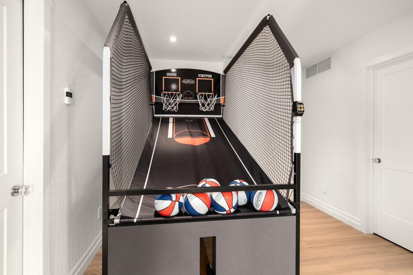 A basketball court with two hoops and a net, and a row of volleyballs on the floor.