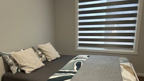 Ein kleines Bett mit grauer Decke und Kissen, ein Fenster mit Jalousien und Blick ins Freie.