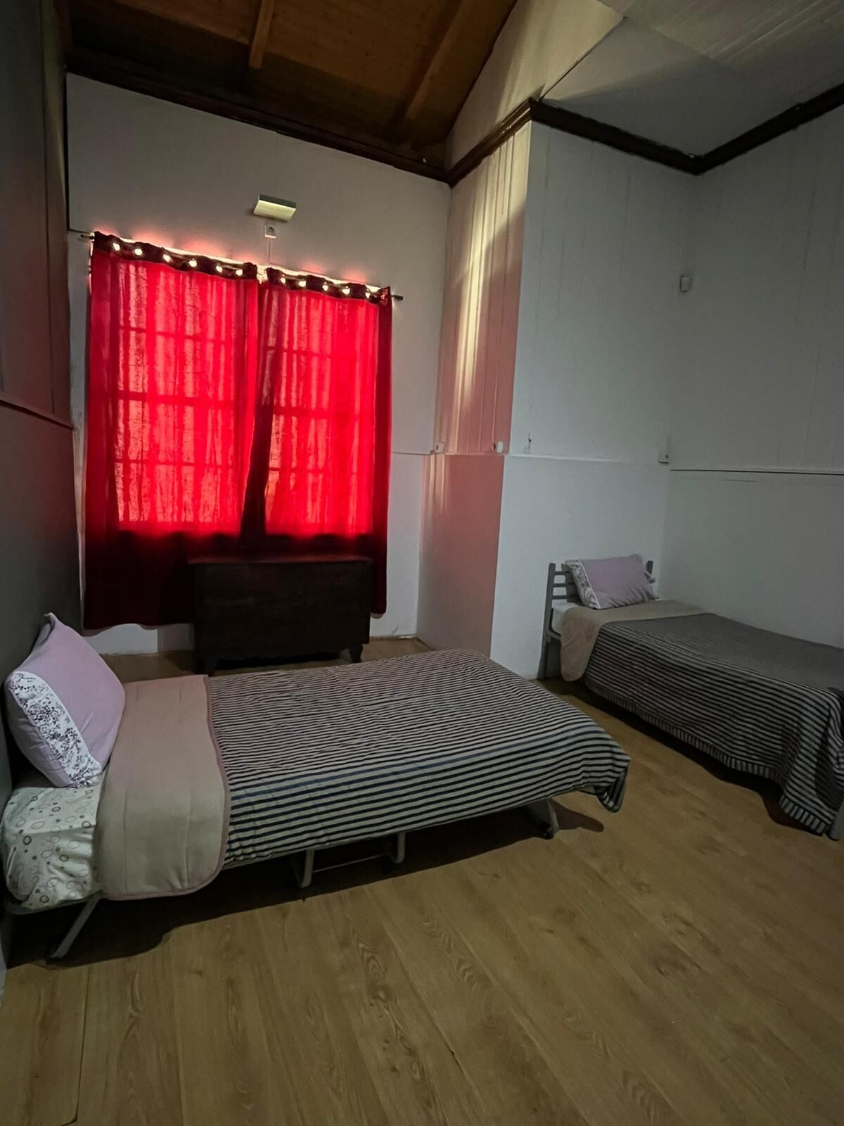 A room with two single beds, a wooden floor, a red curtain, and a small table with a chair.