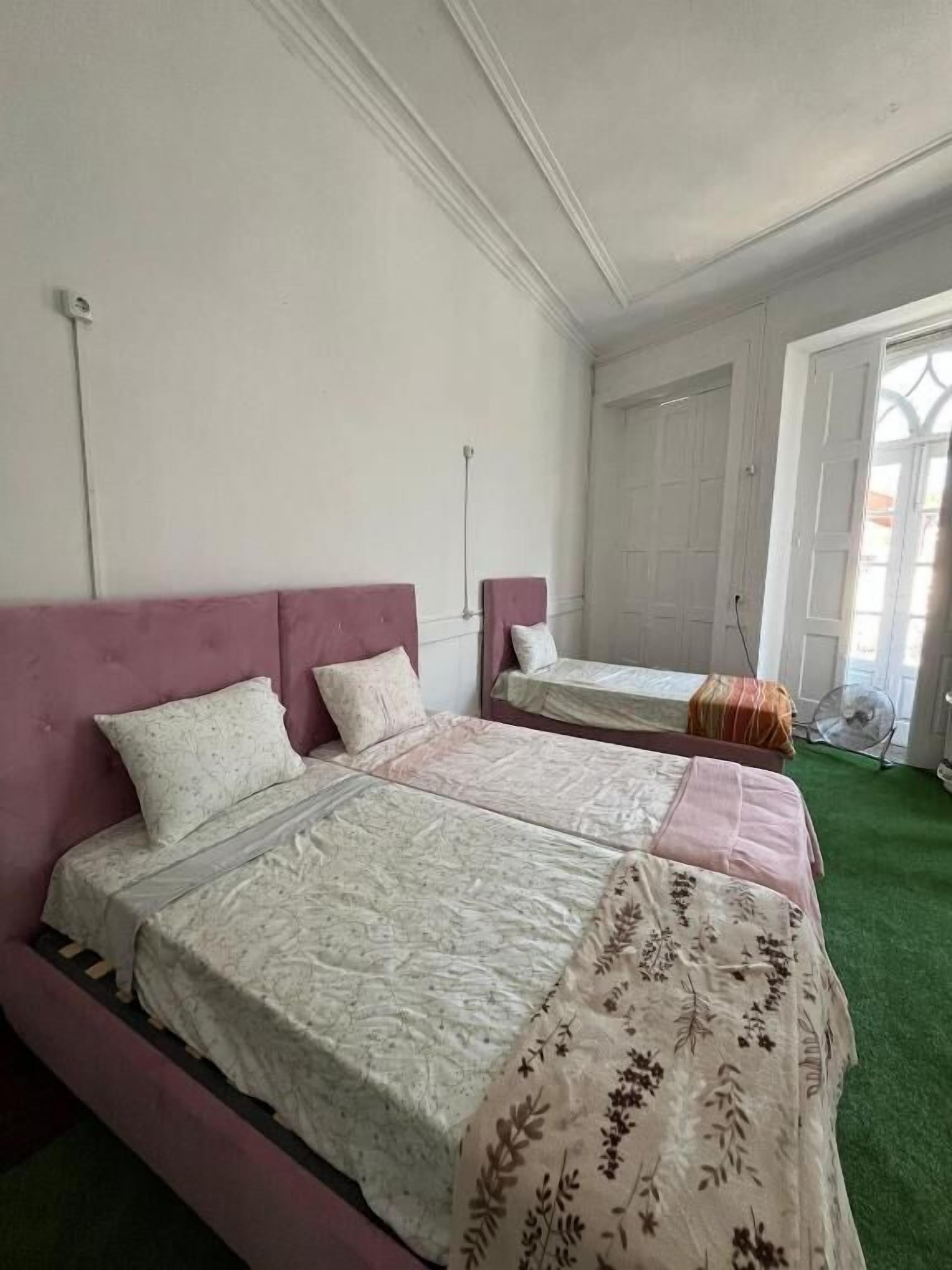 A room with two beds, each with a headboard and pillows, a window with a view, and a door.