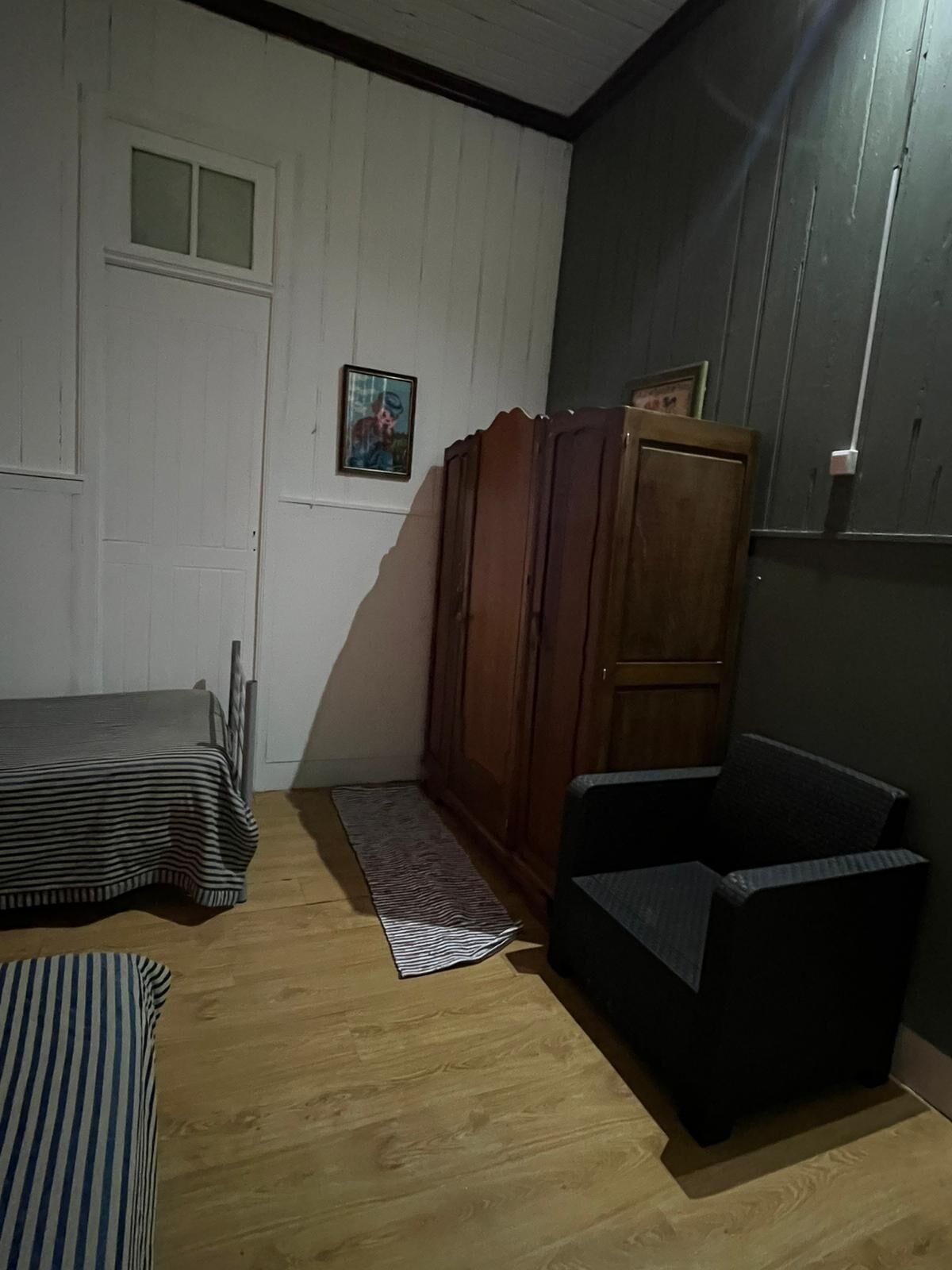 Una habitación pequeña y poco iluminada, con una cama individual, un armario de madera y una silla negra.