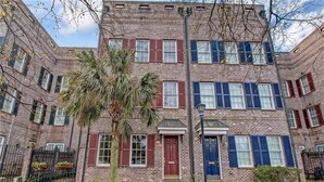 A multi-story brick building with red and blue shutters, a palm tree in the center, and a small garden area. - Historic 3-Bed Townhouse w/ Courtyard & Parking (Savannah)