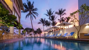 A swimming pool with lounge chairs and umbrellas, surrounded by palm trees and a building with a balcony. - The Lucky Elephant (Hikkaduwa)