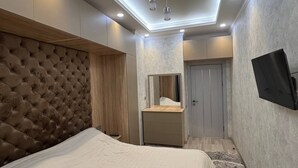 Une chambre à coucher avec une tête de lit capitonnée, une table de chevet, un miroir, une commode et un téléviseur à écran plat fixé au mur.
