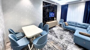 Een moderne woonkamer met een hoekbank, een eettafel met stoelen, een televisie bevestigd aan een zwarte marmeren muur en een groot raam met blauwe gordijnen.