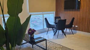 A modern living room with a large window, a flat-screen TV mounted on the wall, a coffee table, and a seating area with two chairs.