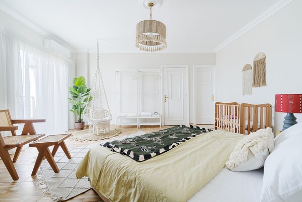 Une chambre à coucher lumineuse et bien éclairée, avec un grand lit, un fauteuil suspendu, un berceau en bois et une plante en pot.