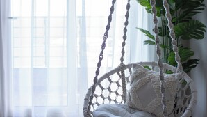 A room with a macrame hanging chair, a large potted plant, and a view through sheer curtains.