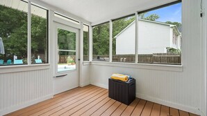 Un porche cubierto con piso de madera y paredes blancas, que cuenta con ventanas amplias y un contenedor negro.