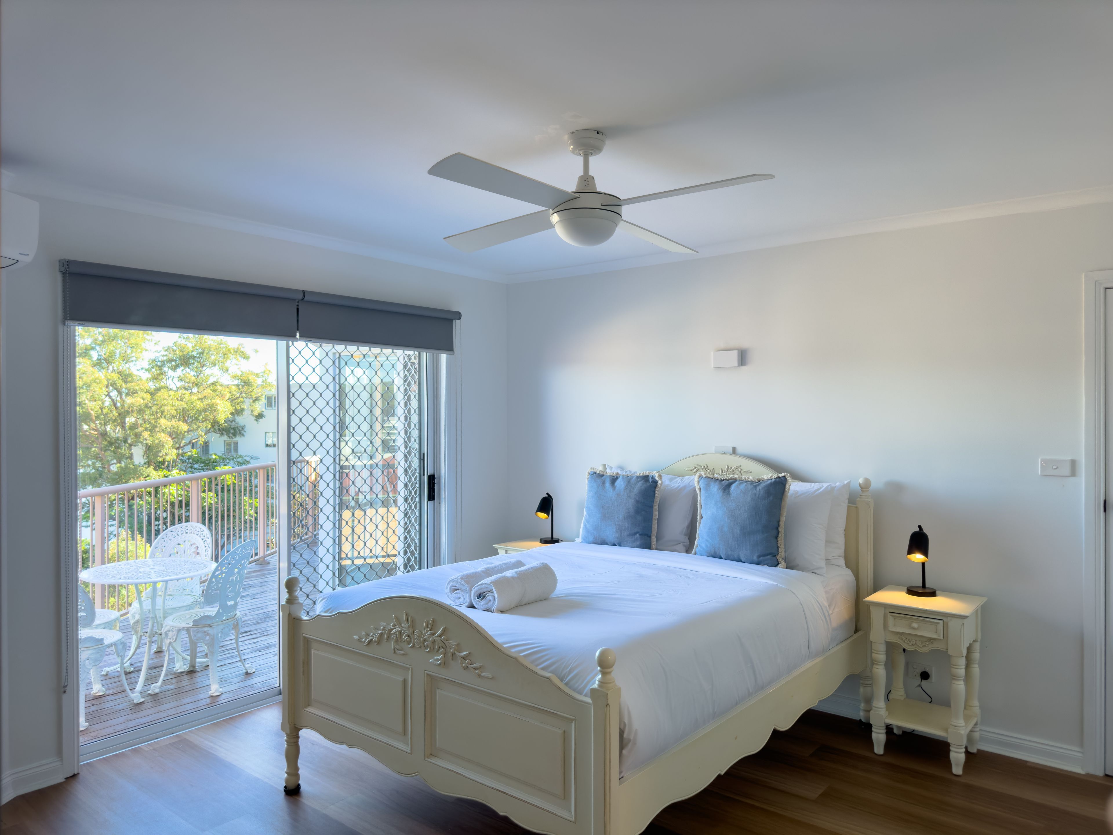 Ein Schlafzimmer mit einem Bett, einem Deckenventilator, einem Nachttisch mit Lampe und einem Balkon mit Stühlen.