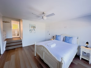 Een slaapkamer met een bed, nachtkastje, bijzettafeltje, plafondventilator en een deur naar de badkamer.