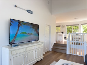 A living room with a television displaying a beach scene, a white cabinet, and a staircase leading to a dining area. - Tomaree Sands Family Townhouse in Nelson Bay (Nelson Bay)
