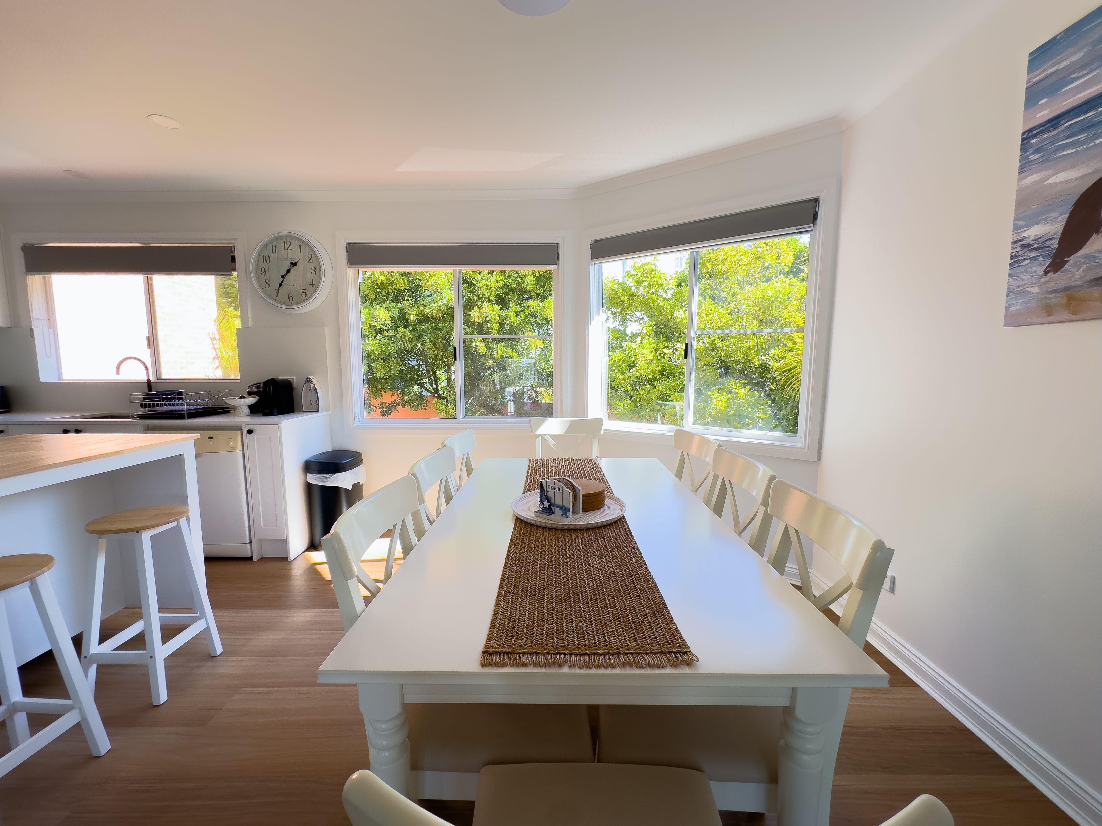 A modern kitchen with white cabinetry, a dining area with a white table, and large windows with a view of greenery.