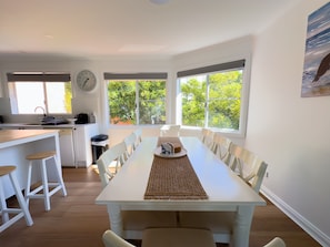 Een moderne keuken met witte kasten, een eethoek met een witte tafel, en grote ramen met uitzicht op groen.